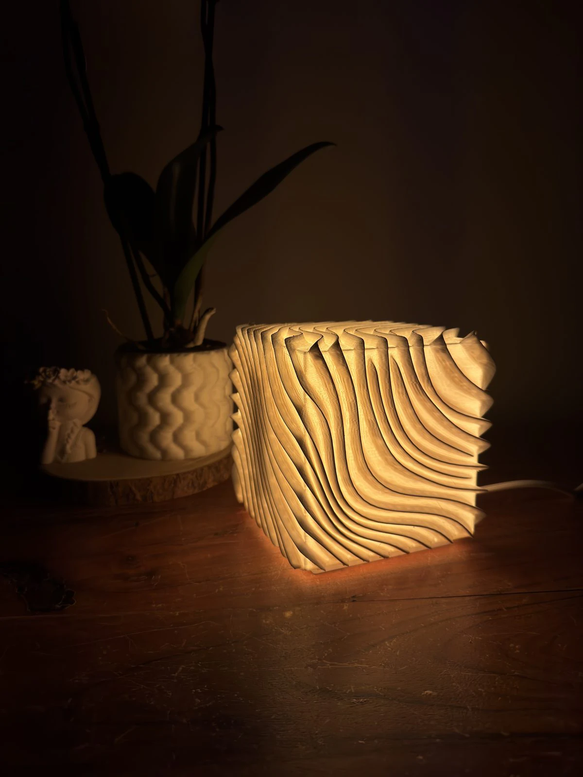 O candeeiro Maré aceso numa mesa de madeira, em casa, ao lado de uma planta